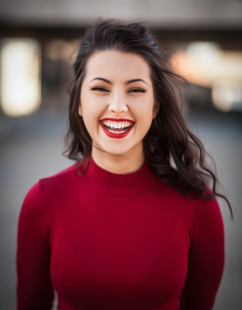 A woman in a red turtleneck laughing with her eyes closed, against a soft grey background.