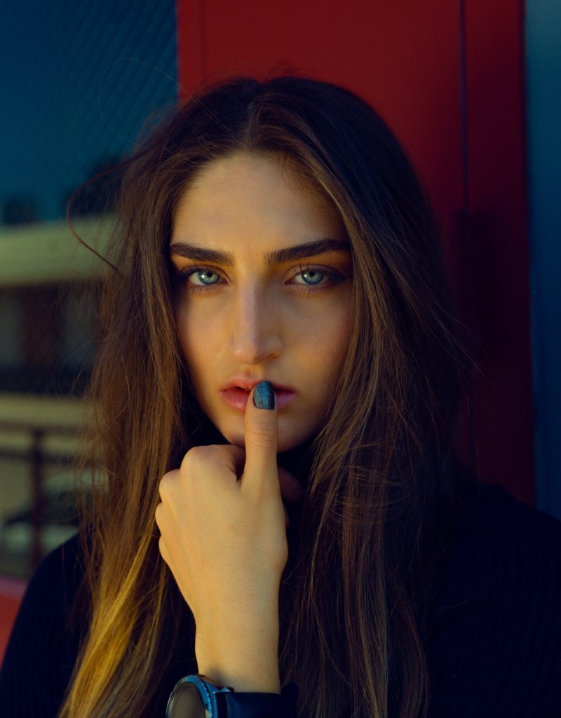 A woman with pale blue eyes holding a finger to her lips, in front of a red and blue wall.